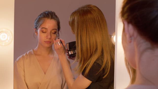 A skilled makeup artist applies eye shades to a young beautiful model in front of the mirror adorned with soft lit bulbs.