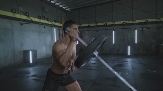 A young muscular man is training his toned physique by lifting the gym bar with just one hand.