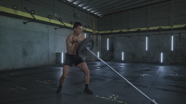 A young shirtless muscular man lifting with just one hand a heavy barbell at the gym.