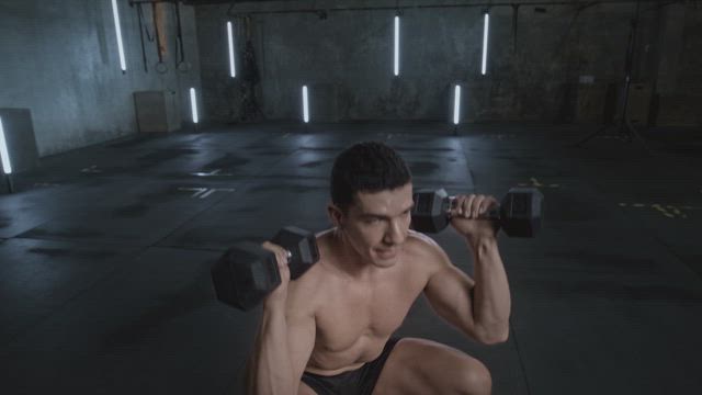 A young shirtless young man shows his physique and toned body by doing a combined exercise with squats and dumbbell lifting.