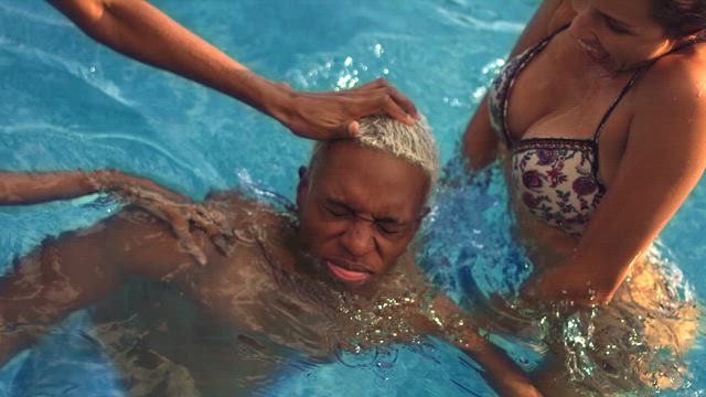 Two young women wearing bikinis push a young black man into the deep refreshing waters of the swimming pool while they laugh.